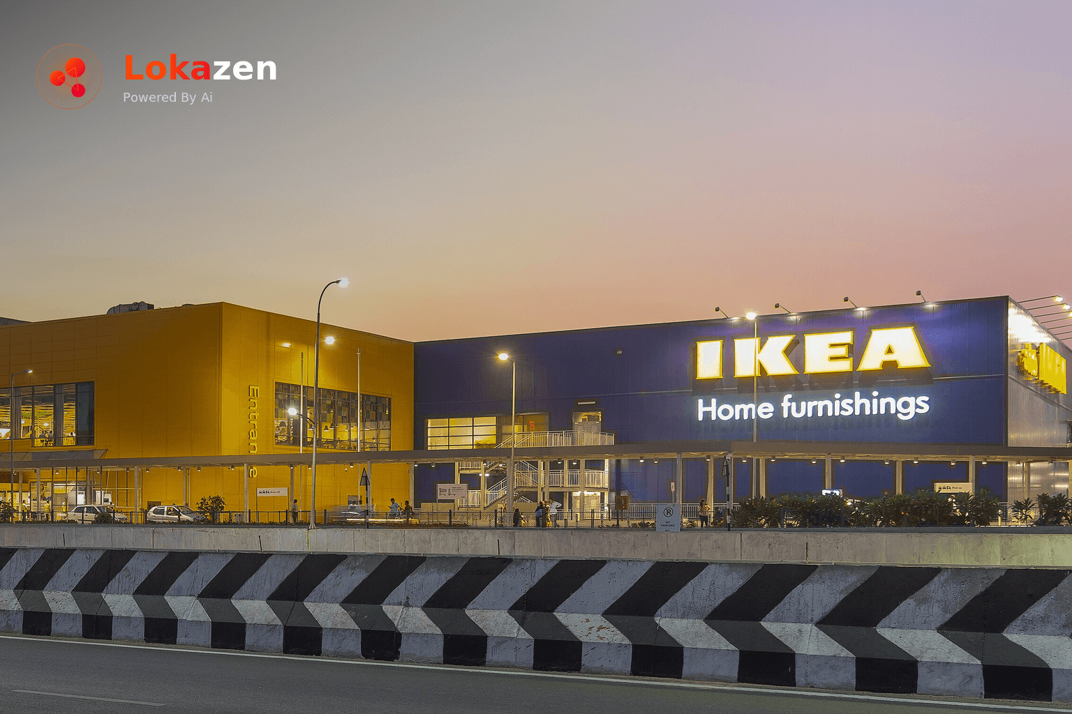 Evening exterior of the IKEA Hyderabad store under a pink and orange sky, with its blue and yellow building, lit IKEA Home furnishings sign and a highway divider in the foreground